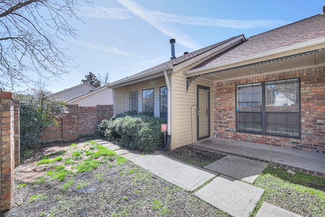 view of exterior entry featuring brick siding, a shingled roof, and fence