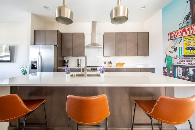 kitchen with dark brown cabinetry, light countertops, wall chimney exhaust hood, stainless steel fridge, and a center island with sink