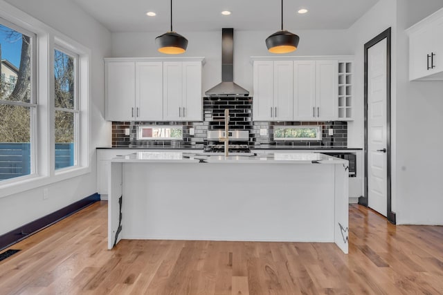 kitchen featuring light wood-type flooring, wall chimney range hood, a kitchen island with sink, and white cabinets