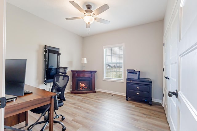 office space with a warm lit fireplace, light wood-style flooring, baseboards, and a ceiling fan