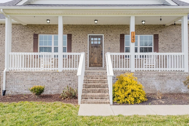 view of exterior entry with covered porch, brick siding, and roof with shingles