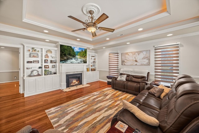 living area featuring ceiling fan, a fireplace with flush hearth, wood finished floors, ornamental molding, and a raised ceiling