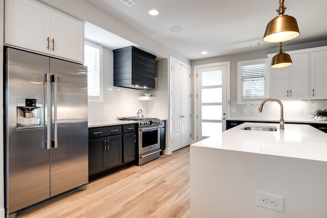 kitchen with appliances with stainless steel finishes, hanging light fixtures, light wood-style floors, premium range hood, and a sink