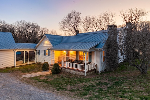 back of house at dusk with driveway, covered porch, metal roof, and a chimney