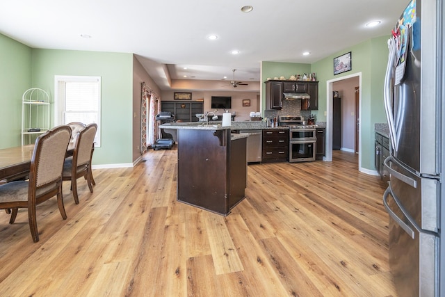 kitchen with light wood finished floors, appliances with stainless steel finishes, a breakfast bar area, dark brown cabinets, and under cabinet range hood