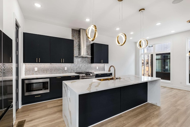 kitchen with light wood finished floors, dark cabinets, stainless steel appliances, wall chimney range hood, and a sink