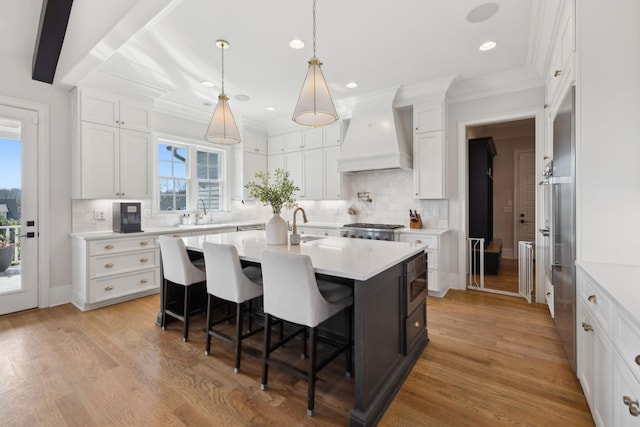 kitchen with custom exhaust hood, light countertops, an island with sink, light wood-type flooring, and a kitchen bar