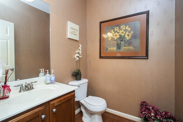 bathroom with toilet, vanity, wood finished floors, baseboards, and wallpapered walls