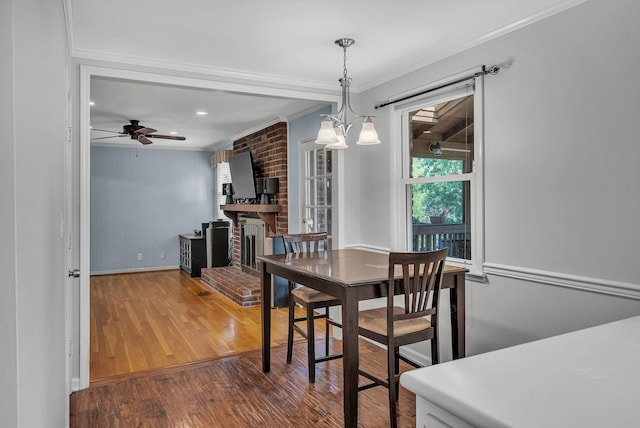 dining space with a fireplace, crown molding, wood finished floors, baseboards, and ceiling fan with notable chandelier