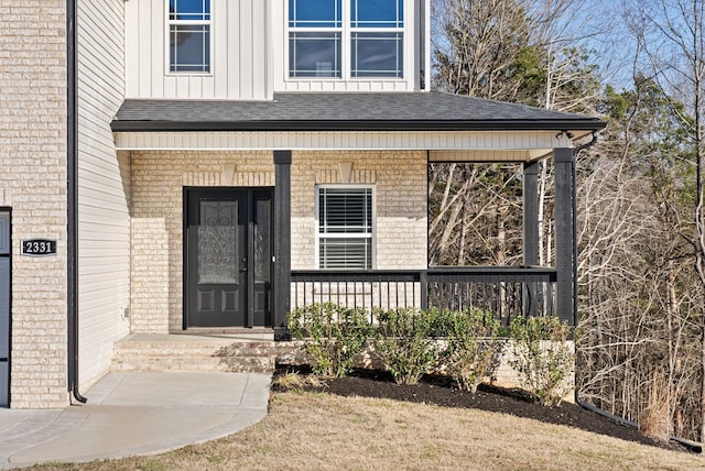 entrance to property featuring roof with shingles, brick siding, board and batten siding, and a porch