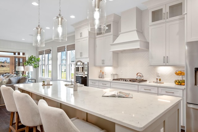 kitchen featuring tasteful backsplash, custom range hood, appliances with stainless steel finishes, open floor plan, and a kitchen island with sink