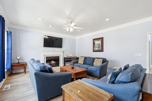 living room featuring a warm lit fireplace, visible vents, a ceiling fan, ornamental molding, and light wood finished floors