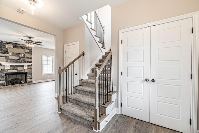 staircase featuring a large fireplace, ceiling fan, baseboards, and wood finished floors