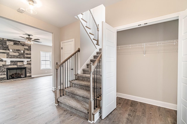 staircase with a stone fireplace, wood finished floors, a ceiling fan, and baseboards