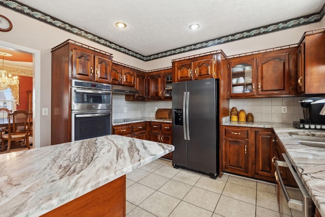 kitchen with light tile patterned floors, under cabinet range hood, stainless steel appliances, light stone countertops, and glass insert cabinets