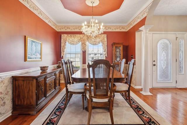 dining room featuring decorative columns, a raised ceiling, hardwood / wood-style flooring, a wainscoted wall, and an inviting chandelier