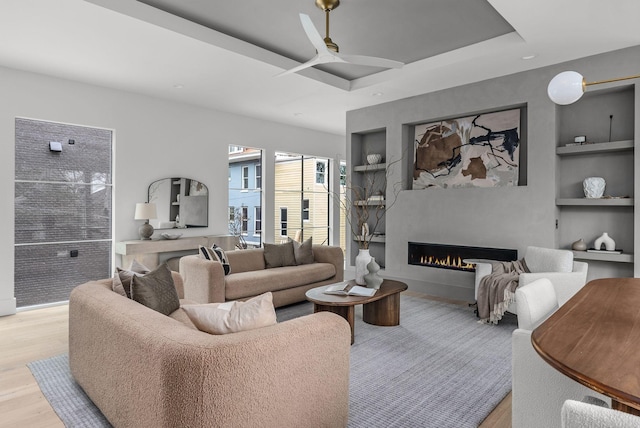 living area featuring ceiling fan, built in shelves, wood finished floors, a lit fireplace, and a raised ceiling