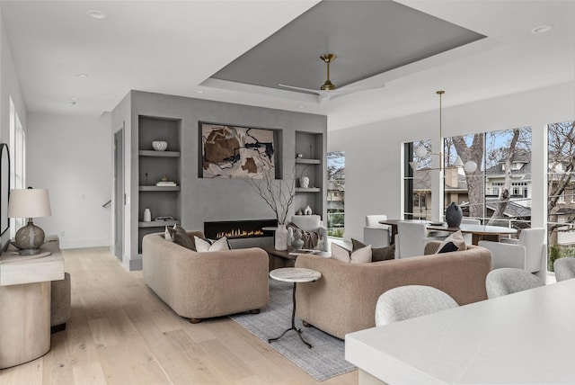 living room with a warm lit fireplace, built in shelves, light wood-style floors, a tray ceiling, and plenty of natural light