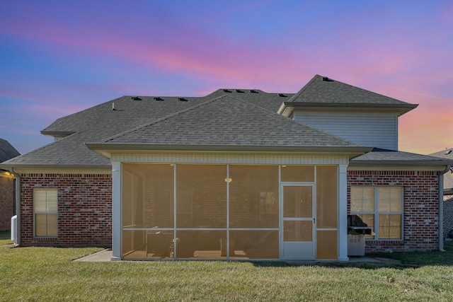 back of house at dusk featuring a yard, brick siding, roof with shingles, and a sunroom