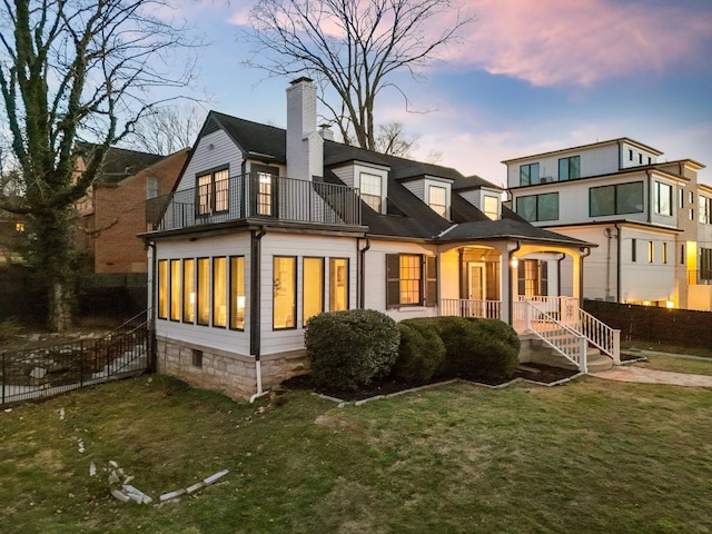 back of property at dusk with a balcony, covered porch, fence, a yard, and a chimney