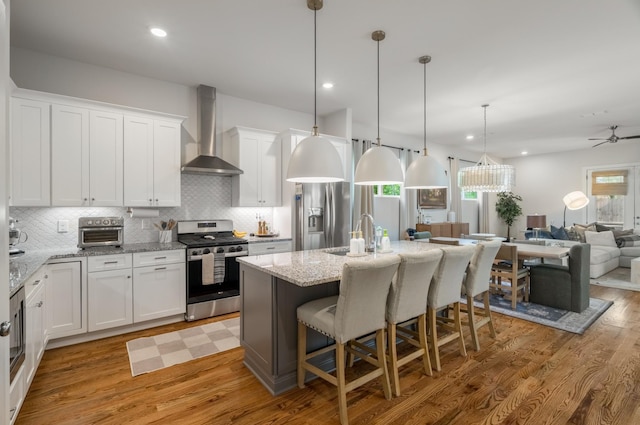 kitchen featuring appliances with stainless steel finishes, open floor plan, white cabinets, a sink, and wall chimney range hood