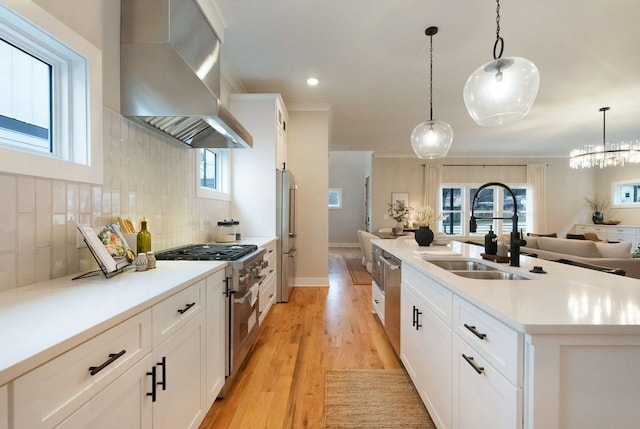 kitchen featuring stainless steel appliances, a sink, wall chimney range hood, light wood-type flooring, and backsplash