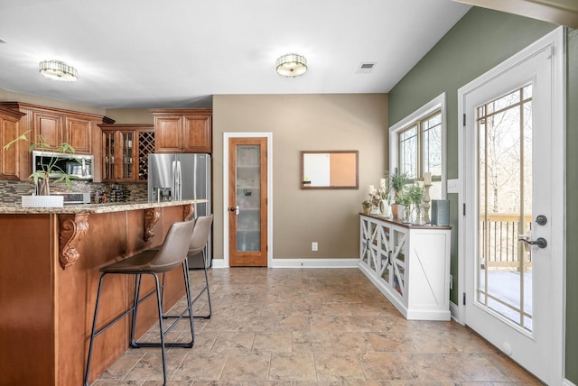 kitchen with backsplash, appliances with stainless steel finishes, brown cabinetry, glass insert cabinets, and a kitchen bar