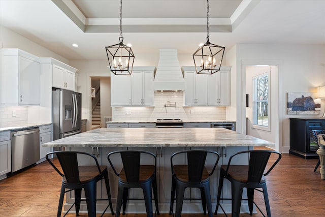 kitchen with dark wood-style flooring, stainless steel appliances, a raised ceiling, a kitchen island, and premium range hood