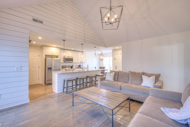 living area with a notable chandelier, light wood finished floors, visible vents, wood walls, and high vaulted ceiling