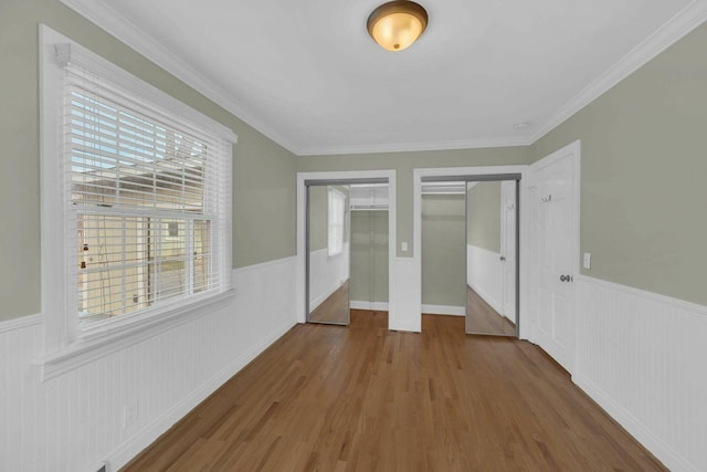 unfurnished bedroom featuring multiple closets, a wainscoted wall, ornamental molding, and wood finished floors