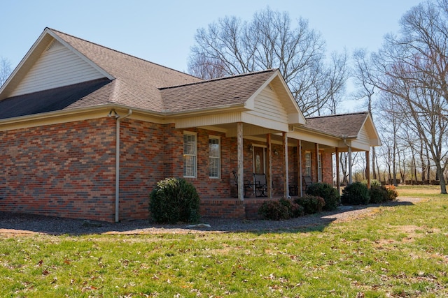 view of side of property featuring covered porch, roof with shingles, brick siding, and a lawn