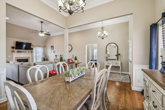 dining area with crown molding, dark wood finished floors, lofted ceiling, a lit fireplace, and ceiling fan with notable chandelier