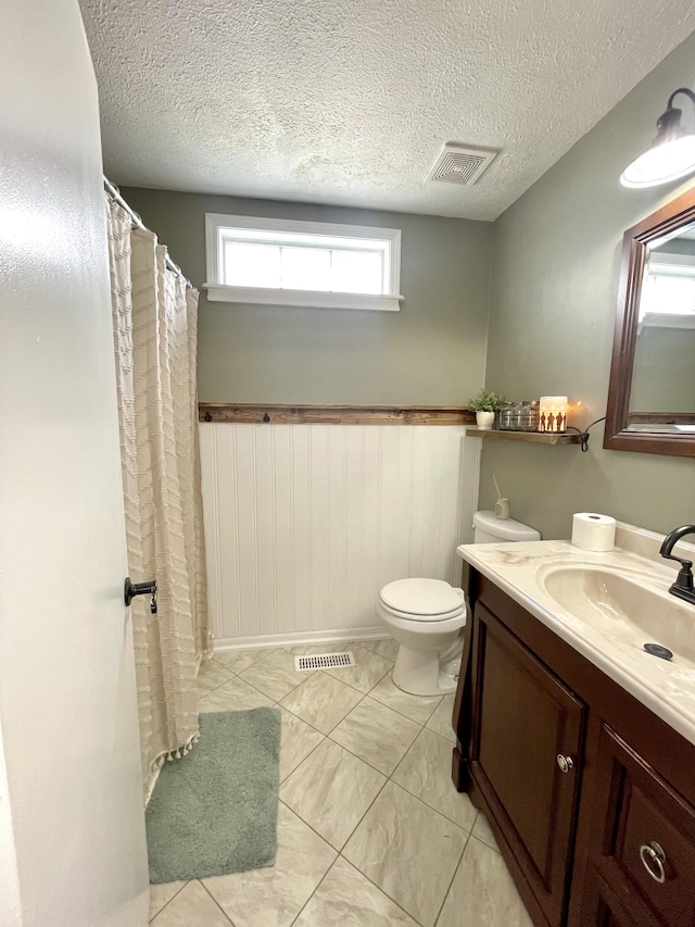 bathroom with a textured ceiling, toilet, vanity, visible vents, and wainscoting