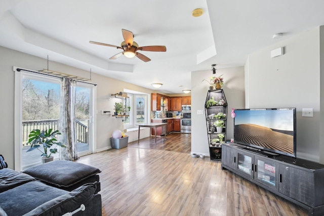 living area featuring light wood-style floors, a tray ceiling, ceiling fan, and baseboards