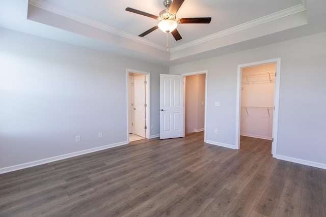 unfurnished bedroom featuring baseboards, a raised ceiling, ornamental molding, dark wood-type flooring, and a walk in closet