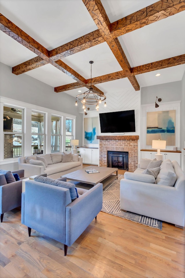 living room with beam ceiling, an inviting chandelier, a brick fireplace, light wood-type flooring, and coffered ceiling