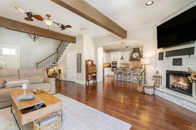 living area featuring stairway, a stone fireplace, wood finished floors, beamed ceiling, and baseboards