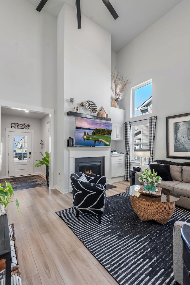 living room featuring light wood-style flooring, a high ceiling, ceiling fan, a lit fireplace, and baseboards