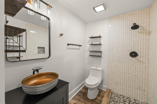 bathroom with toilet, vanity, wood finished floors, tiled shower, and baseboards