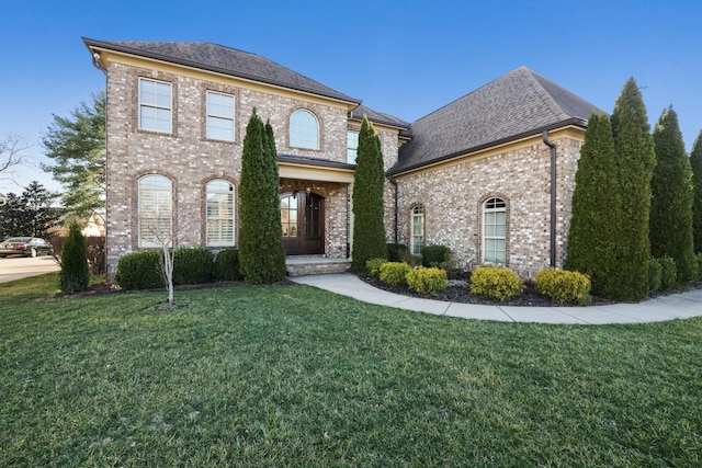 french country inspired facade featuring a shingled roof, a front yard, and brick siding