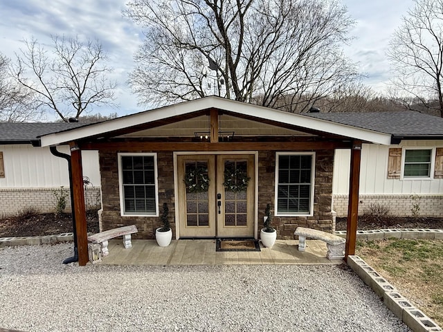 view of exterior entry with a shingled roof, stone siding, covered porch, french doors, and board and batten siding