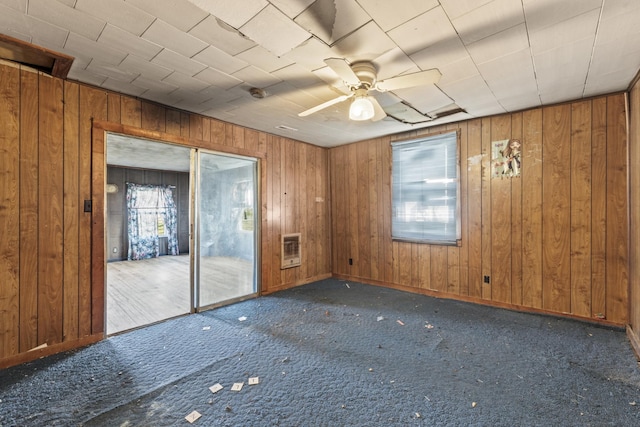 carpeted spare room featuring baseboards, wooden walls, a ceiling fan, and heating unit