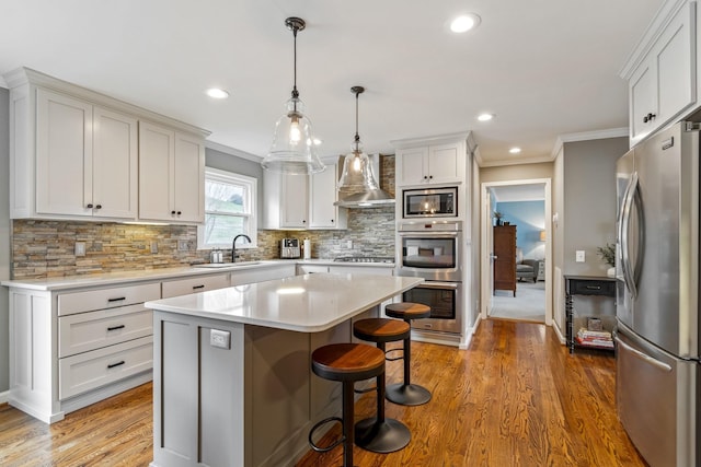 kitchen featuring a breakfast bar area, a sink, light countertops, appliances with stainless steel finishes, and wall chimney exhaust hood