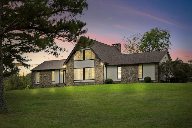 view of front of property with a shingled roof, stone siding, a lawn, and a chimney