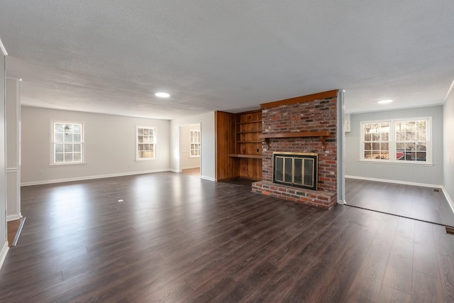 unfurnished living room with a brick fireplace, built in shelves, baseboards, and dark wood finished floors