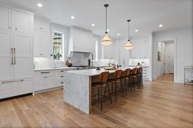 kitchen featuring light wood-style floors, white cabinets, custom range hood, and an island with sink