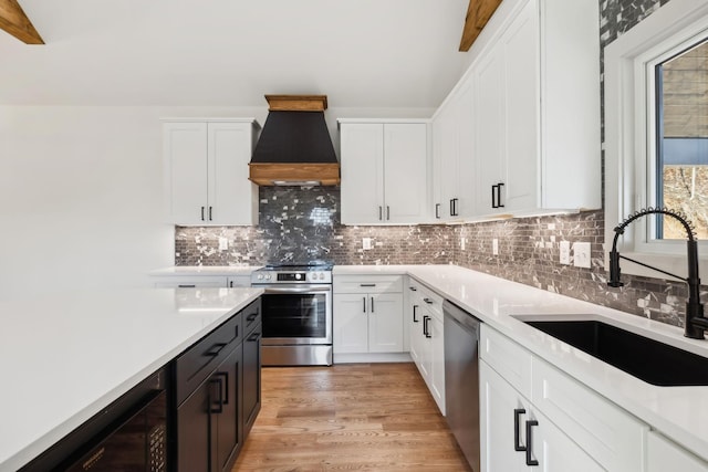 kitchen featuring white cabinets, wine cooler, custom range hood, appliances with stainless steel finishes, and a sink