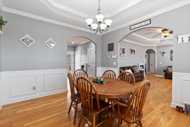 dining room with arched walkways, a raised ceiling, wainscoting, light wood-type flooring, and ceiling fan with notable chandelier