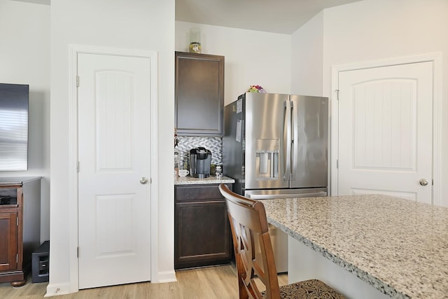 kitchen featuring dark brown cabinetry, stainless steel fridge with ice dispenser, light wood-style flooring, light stone countertops, and backsplash