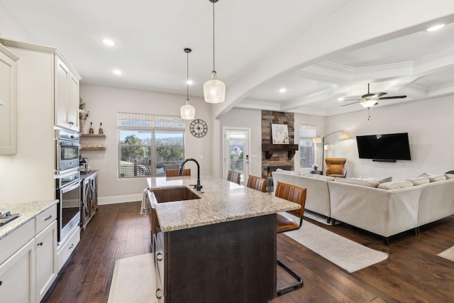 kitchen with a breakfast bar, dark wood-style flooring, a kitchen island with sink, a sink, and a stone fireplace